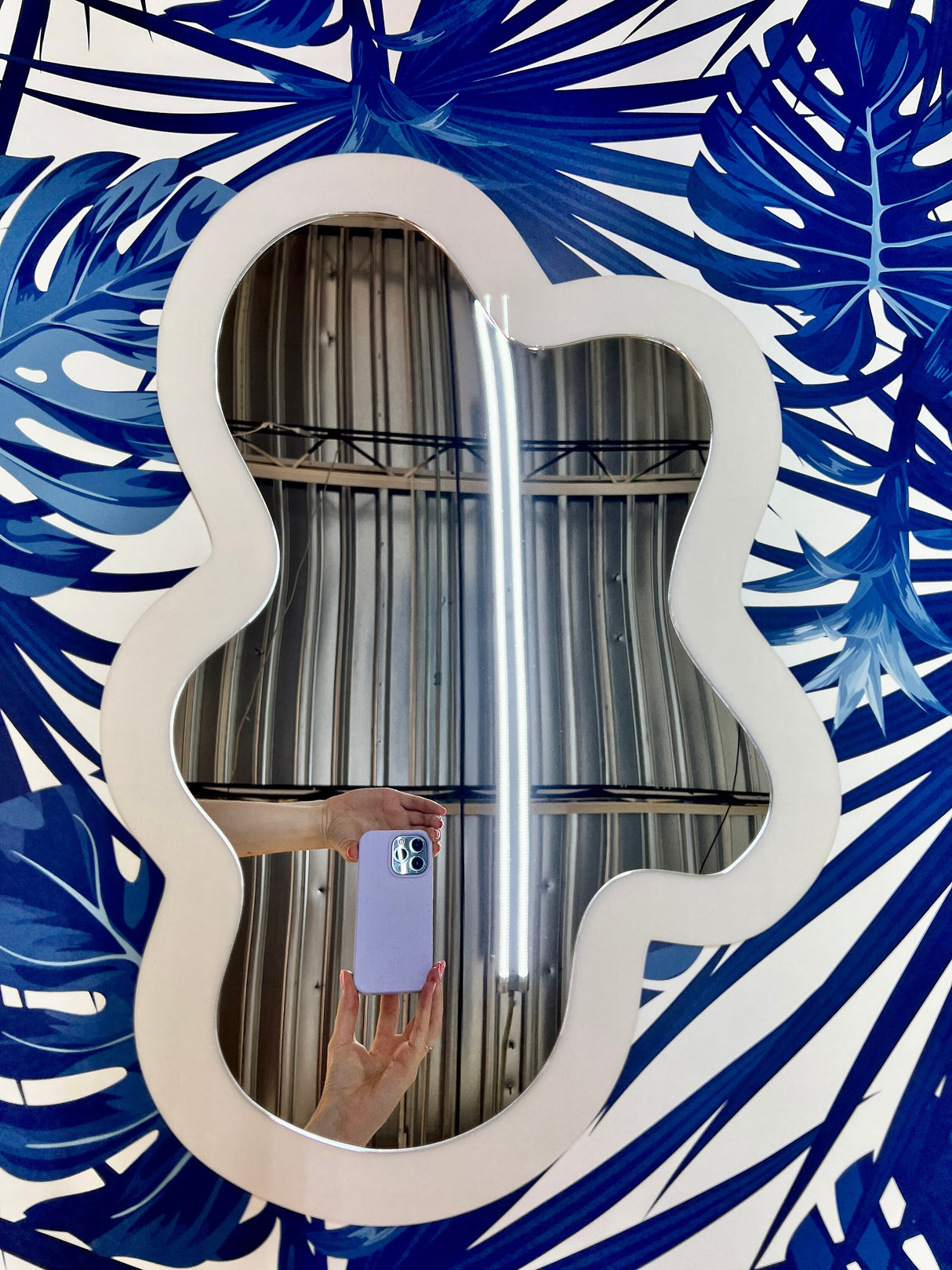 The uniQstiQ Irregular Mirror (Acrylic Art, Asymmetrical/Blob Mirrored, Modern Wall Décor) reflects a hand holding a purple phone over a blue leaf-patterned surface. Custom colors available.