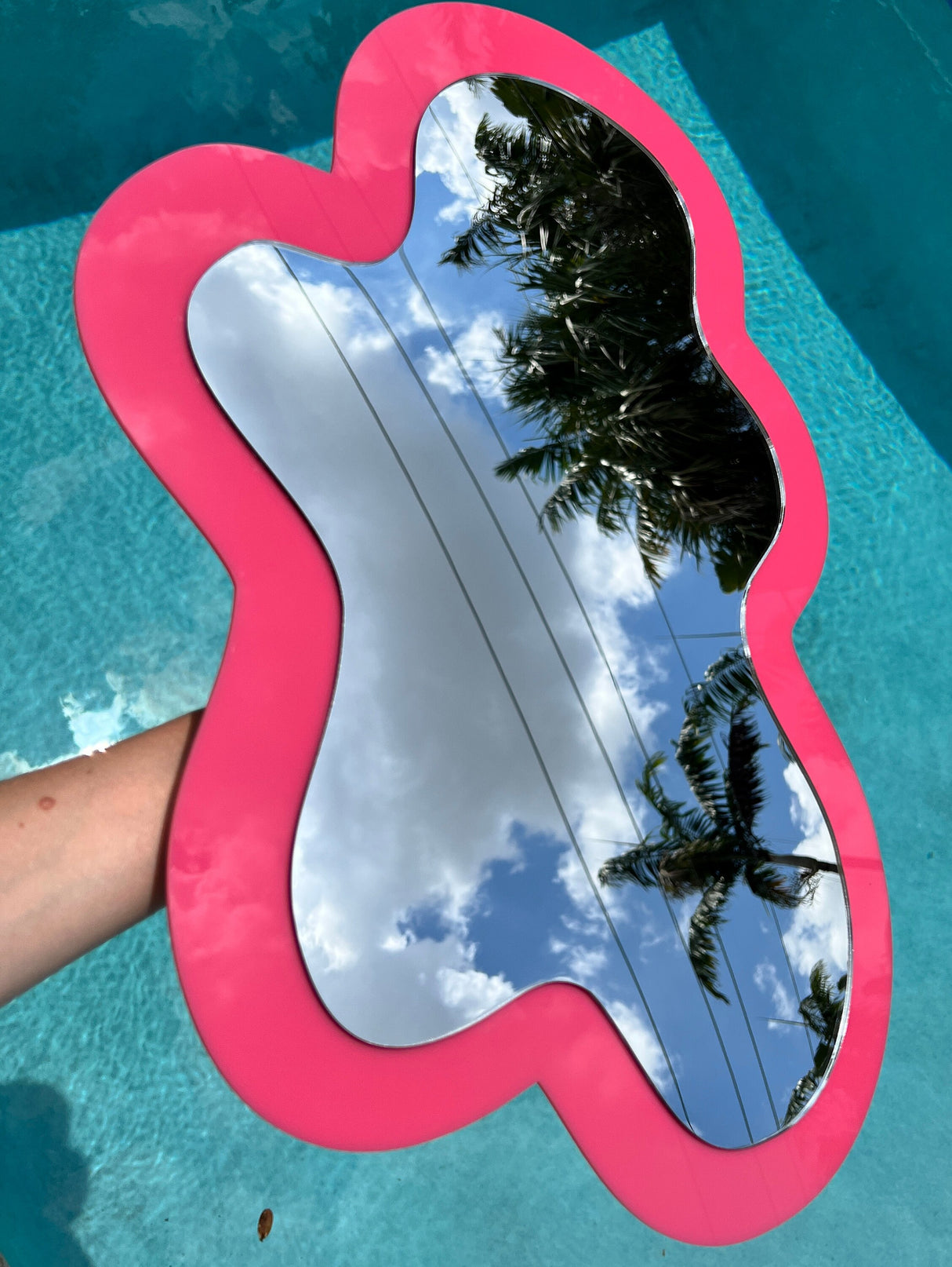 The Irregular Mirror by uniQstiQ features a custom pink acrylic design, reflecting clouds, palm trees, and power lines over a blue pool—perfect as asymmetrical modern art or unique abstract wall decor.