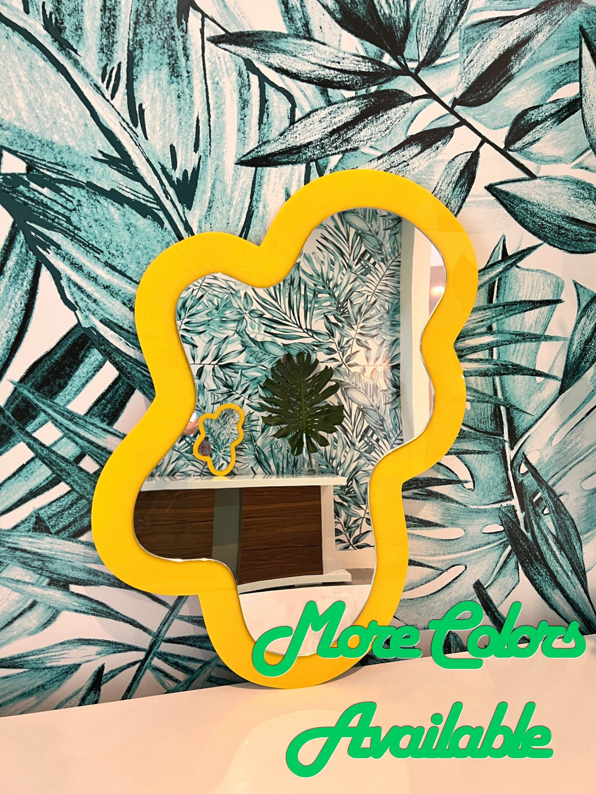 The uniQstiQ Irregular Mirror features an asymmetrical acrylic blob design on leafy wallpaper, inspired by modern pop art decor. More Colors Available is shown in green at the bottom.